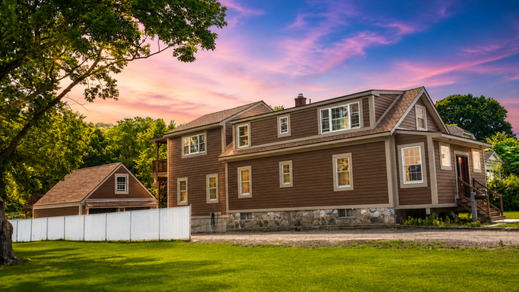 Custom-built two-story home surrounded by mature trees and lush landscaping, featuring brown siding, stone foundation accents, and a detached garage. The property is showcased during a vibrant sunset with pink and blue skies, highlighting the craftsmanship, architectural details, and spacious outdoor setting. Built by Custom Building Professionals LLC, specializing in custom homes and luxury residential construction.